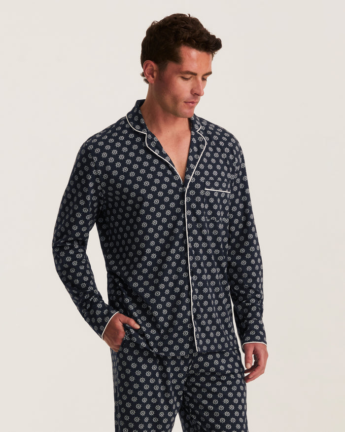 A man wearing the Men’s Lightweight Pima Pajama Set in Captain’s Compass, featuring a navy blue pattern with white piping, stands against a plain light background, looking down with one hand in his pocket.