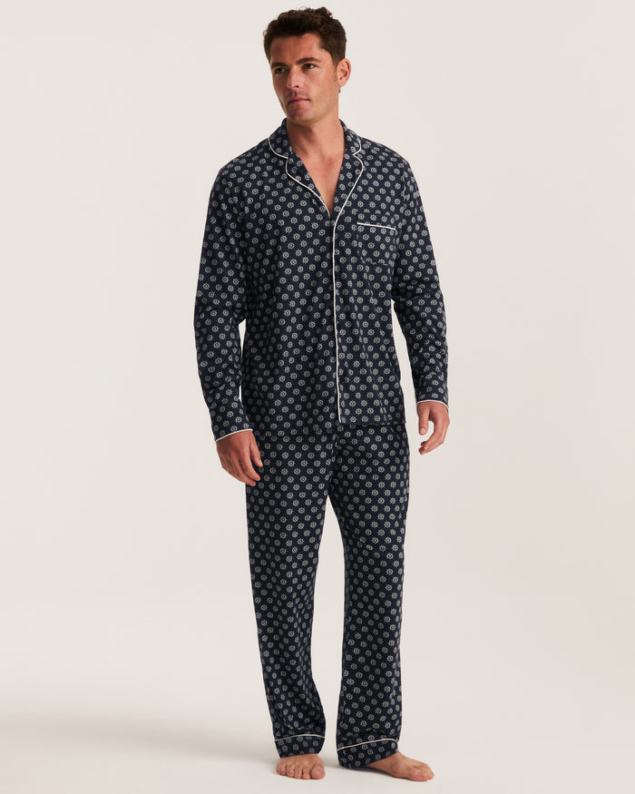 A man stands barefoot wearing the Men’s Lightweight Pima Pajama Set in Captain’s Compass, a matching dark blue pajama set adorned with a white geometric pattern and white piping, featuring a button-up shirt with a chest pocket and long pants.