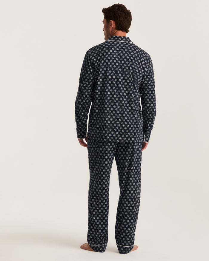 A man stands barefoot facing away, wearing the Men’s Lightweight Pima Pajama Set in Captain’s Compass—a matching navy blue set adorned with a white geometric pattern—against a plain light background.