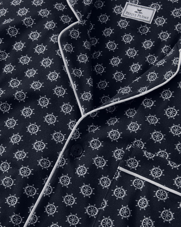 Close-up of the Men’s Lightweight Pima Pajama Set in Captain’s Compass, featuring a navy blue pajama top with a white ship wheel pattern, contrast white piping along the edges, a button closure, a chest pocket, and a Petite Plume label in the upper right corner.