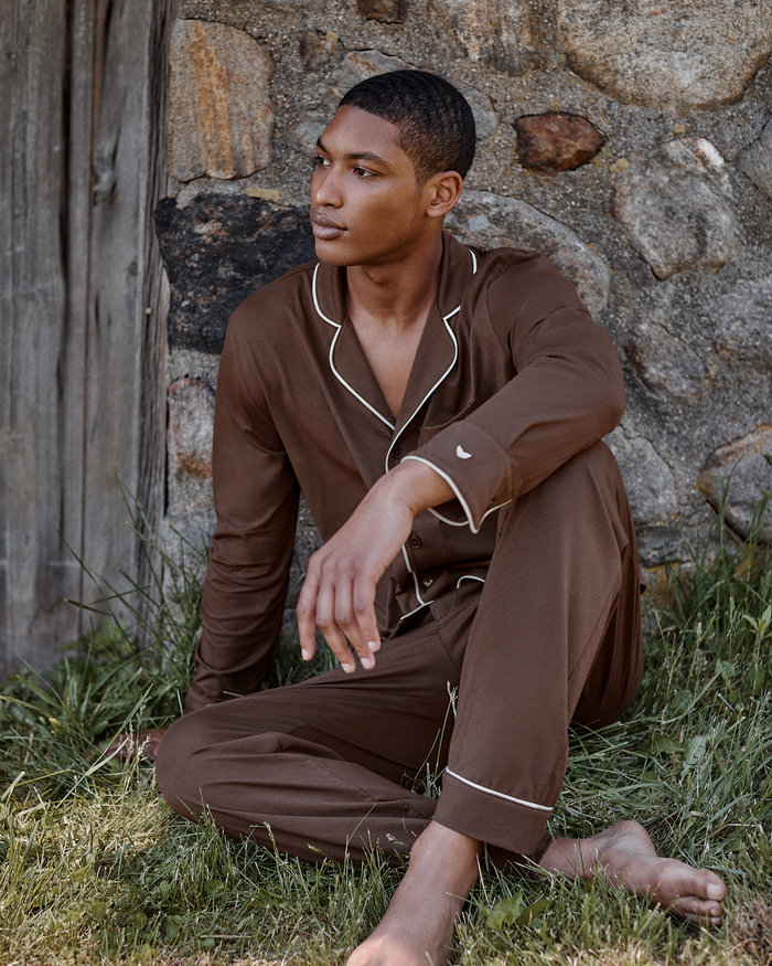 A man wearing the Men’s Lightweight Pima Pajama Set in Espresso sits barefoot on the grass, leaning against a stone wall and wooden door, gazing thoughtfully into the distance.