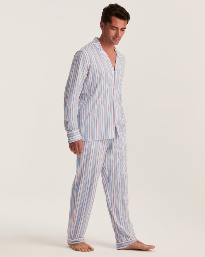 A man stands barefoot against a plain, light background, wearing the Men’s Lightweight Pima Pajama Set in Vintage French Stripes—a light-colored ensemble with vertical blue, pink, and white stripes. He looks down slightly with one hand in his pocket.
