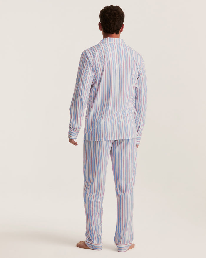 A person stands barefoot facing away, wearing the Men’s Lightweight Pima Pajama Set in Vintage French Stripes—a long-sleeved, button-up design featuring vertical blue, white, and red stripes—against a plain light background.