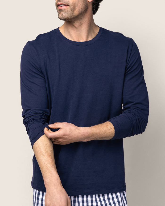 A man adjusts his sleeve, wearing the Petite Plume Mens Pima Crew Neck Long Sleeve Top in Navy with checkered pants, resembling luxurious sleepwear. The background is plain and neutral.