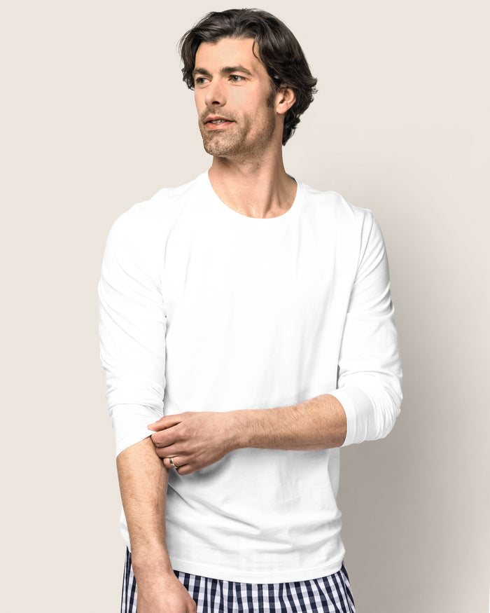 A man wearing a Mens Pima Crew Neck Long Sleeve Top in White by Petite Plume stands against a light background. He gazes left, adjusting one sleeve with his right hand, evoking the calm of a bedtime ritual while paired with checkered pants.