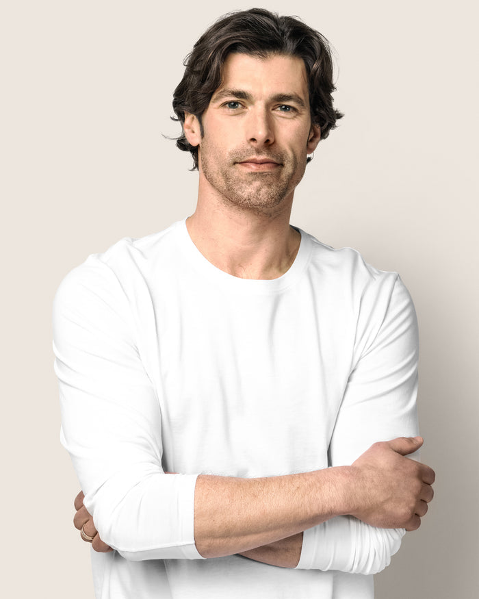 A man with dark, wavy hair and light stubble stands against a plain background. Wearing Petite Plumes Mens Pima Crew Neck Long Sleeve Top in White, his arms are crossed as he looks directly at the camera with a neutral expression, seemingly contemplating his bedtime ritual.
