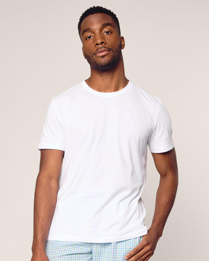 A man wearing a plain white Mens Pima Crew Neck Short Sleeve Top by Petite Plume and light-colored pants stands against a neutral background, looking at the camera with a relaxed expression. His right hand rests gently on his hip, exuding comfort akin to jersey knit pajamas.