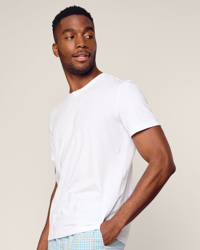 A man in a white Petite Plume Mens Pima Crew Neck Short Sleeve Top and plaid shorts gazes sideways against a neutral background, embodying the effortless comfort typical of luxurious pajamas.
