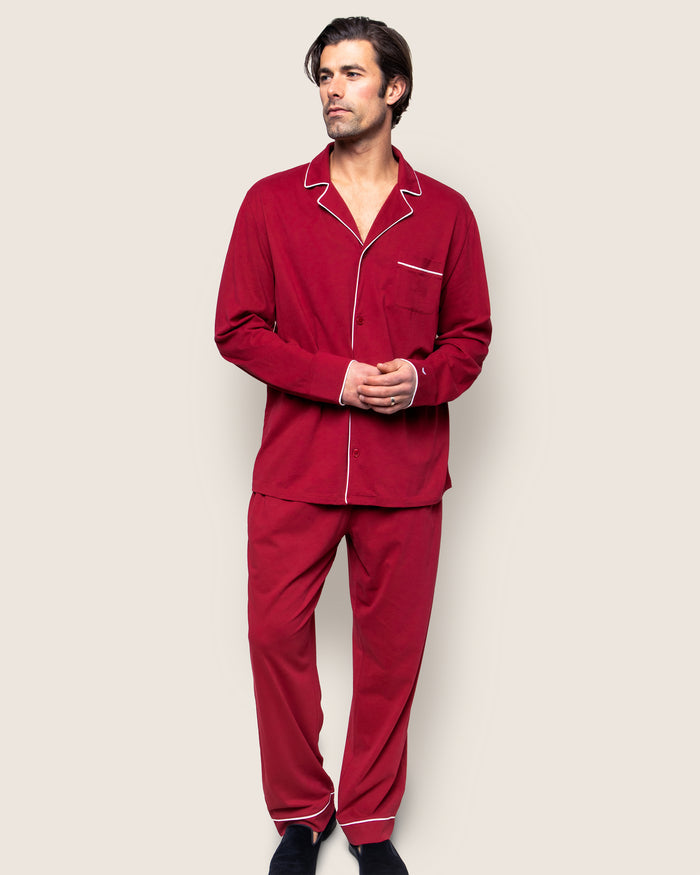 A person in the Mens Pima Pajama Set in Bordeaux by Petite Plume, made from luxurious Peruvian Pima cotton with white piping, stands against a light background. With dark hair and a sideways glance, they have hands clasped in front, wearing the button-up shirt and relaxed pants ensemble.