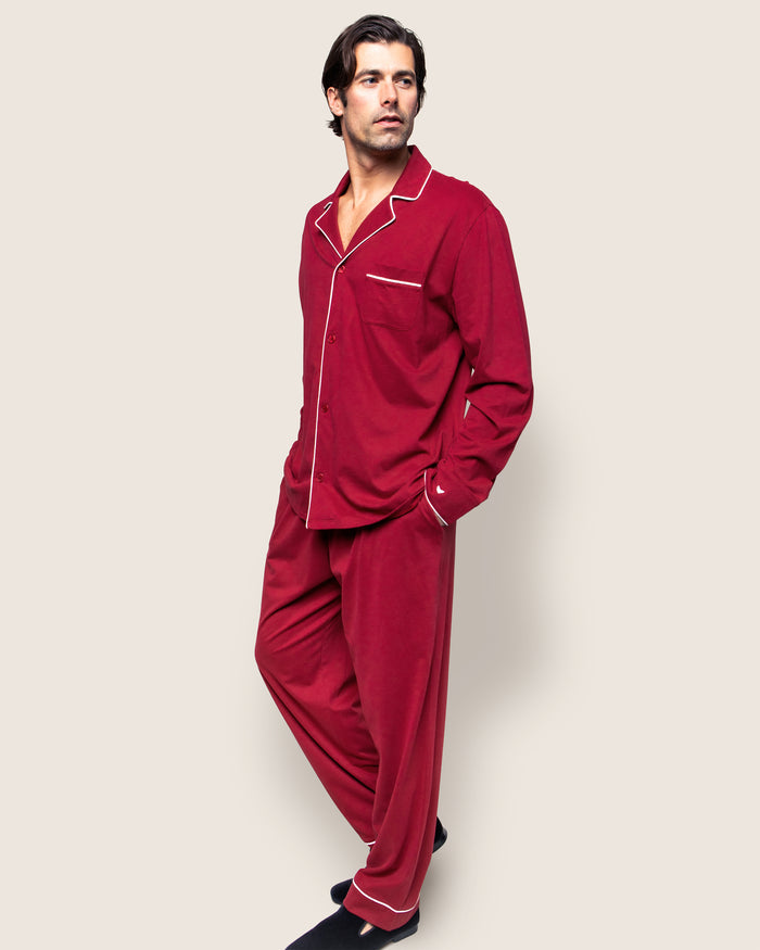 A man in a Mens Pima Pajama Set in Bordeaux by Petite Plume, crafted from luxurious Peruvian Pima cotton, stands with hands in pockets. The timeless set includes a long-sleeved button-up top and matching pants against a plain, light-colored background.