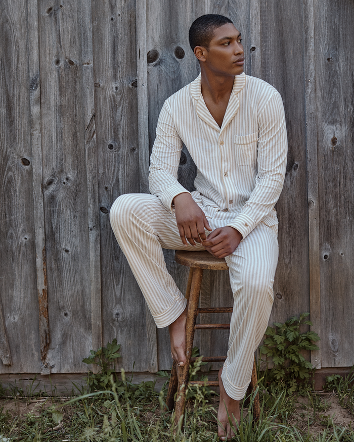 A man wearing the Men’s Pima Pajama Set in Camel Ticking sits barefoot on a wooden stool against a rustic wooden fence, looking to the side with one leg bent and the other touching the ground.