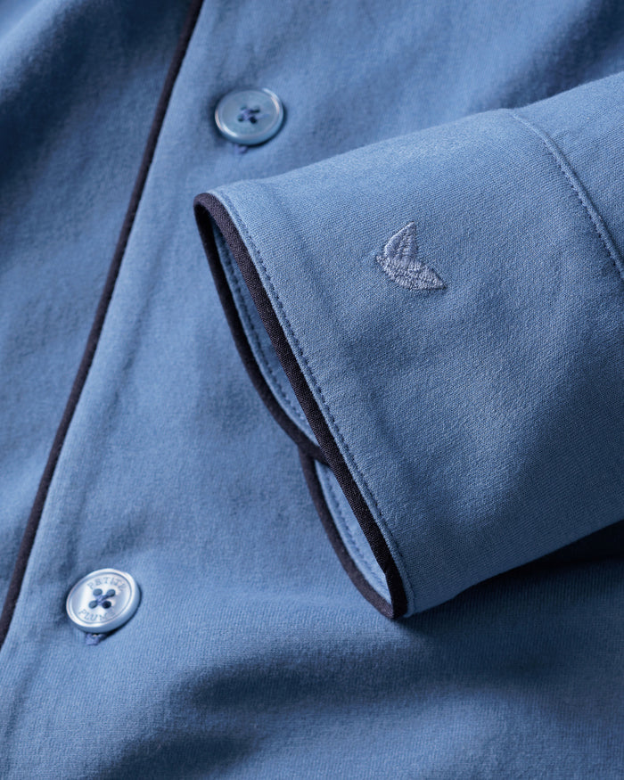 Close-up of Petite Plumes Mens Pima Pajama Set in Mystique Blue features a folded sleeve with an embroidered logo, subtly patterned buttons, and dark fabric piping, embodying luxury sleepwear.