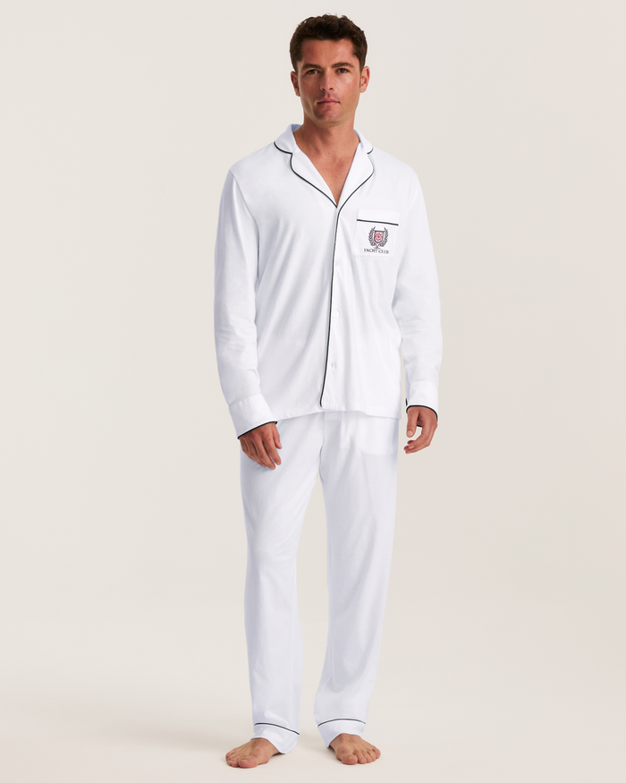 A man stands barefoot in the Men’s Pima Pajama Set in White with Yacht Club Embroidery, featuring black piping and a chest logo, gazing forward with a neutral expression against a plain light background.