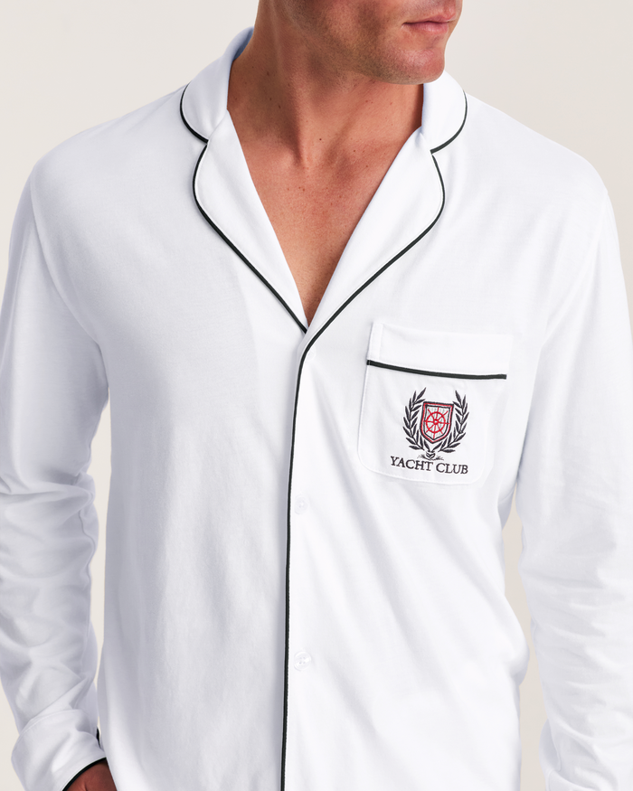 A man wearing the Men’s Pima Pajama Set in White with Yacht Club embroidery, featuring black piping and a crest on the breast pocket, looking to the side.