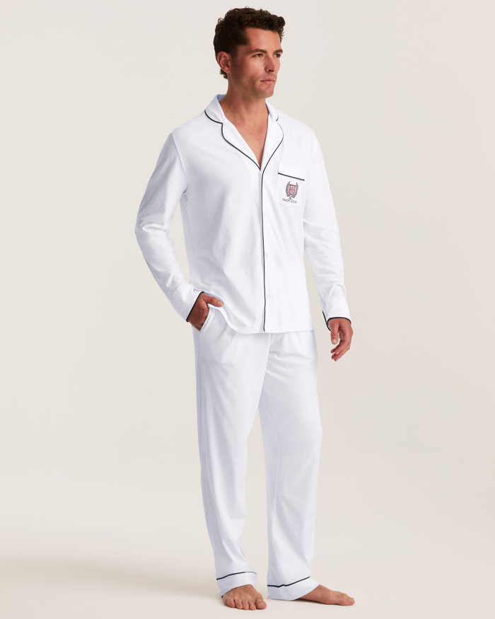 A man stands barefoot against a plain background, wearing the Men’s Pima Pajama Set in White with Yacht Club Embroidery, featuring black piping and a chest pocket as he looks slightly to the side.