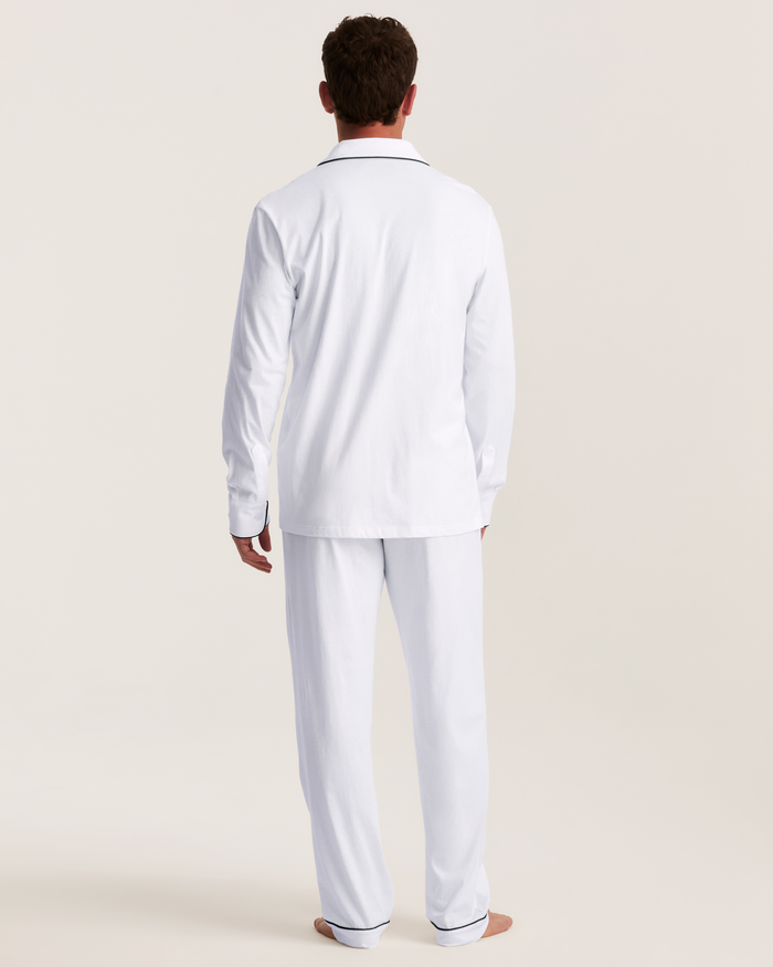 A person stands barefoot, facing away, against a plain light background, wearing the Men’s Pima Pajama Set in White with Yacht Club embroidery, featuring black trim on the cuffs and collar.
