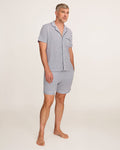 A man stands barefoot in a studio wearing the Men's Pima Pajama Short Set in Navy French Ticking, which features vertical blue and white stripes. He looks slightly to the side with one hand in his pocket.