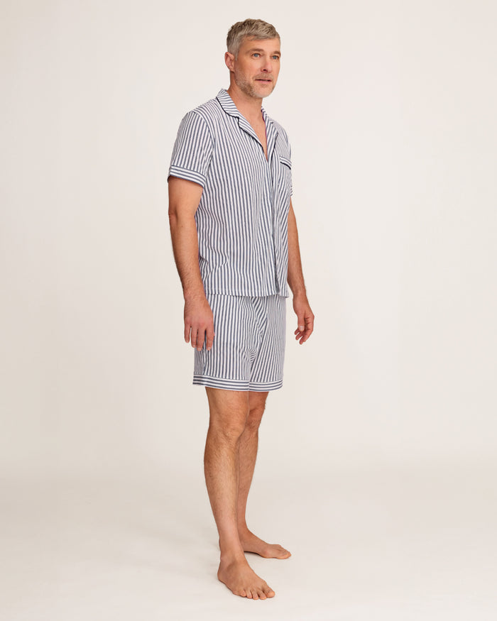 A man stands barefoot in a studio wearing the Men's Pima Pajama Short Set in Navy French Ticking, which features vertical blue and white stripes. He looks slightly to the side with one hand in his pocket.