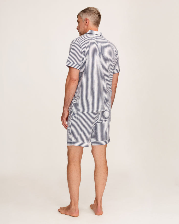 A man stands barefoot in a studio wearing the Men's Pima Pajama Short Set in Navy French Ticking, which features vertical blue and white stripes. He looks slightly to the side with one hand in his pocket.