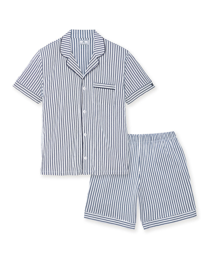 A man stands barefoot in a studio wearing the Men's Pima Pajama Short Set in Navy French Ticking, which features vertical blue and white stripes. He looks slightly to the side with one hand in his pocket.