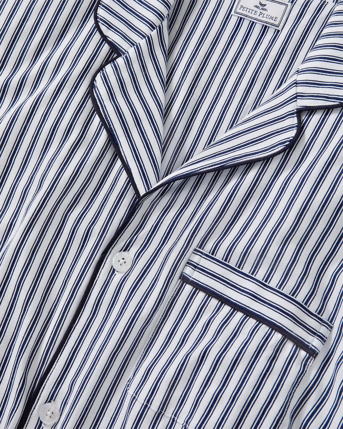 A man stands barefoot in a studio wearing the Men's Pima Pajama Short Set in Navy French Ticking, which features vertical blue and white stripes. He looks slightly to the side with one hand in his pocket.