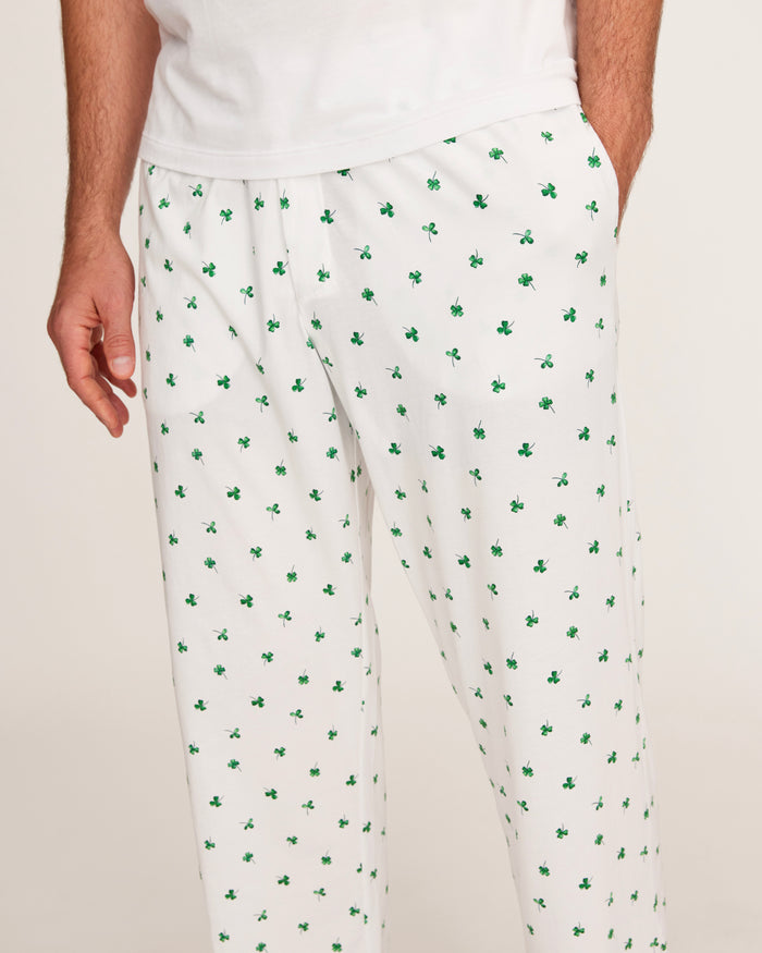 A man with short gray hair stands barefoot against a plain light background, wearing a plain white tee and Men's Pima Pants in Shamrocks—a white pajama pant with a small green pattern.