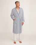 A man stands barefoot in white pajamas and the Men's Pima Robe in Navy French Ticking, belted, looking to the side against a plain light background.