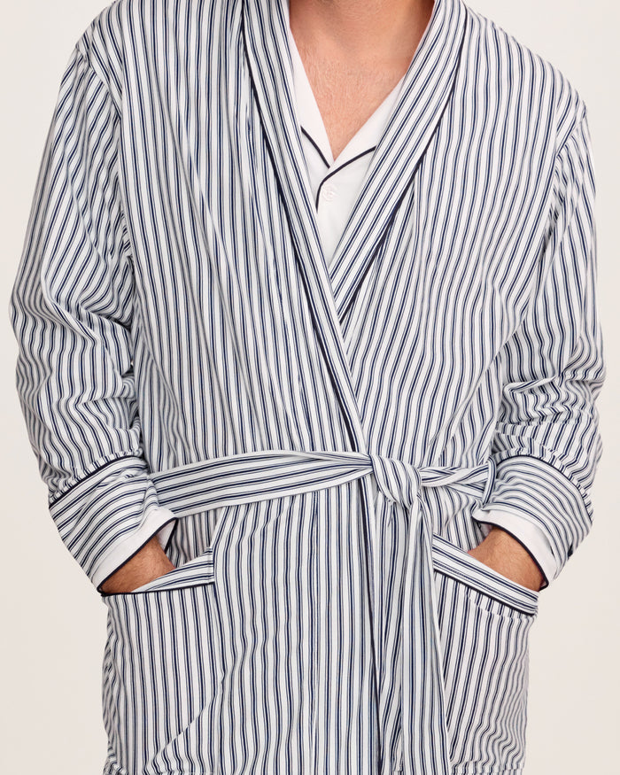 A man stands barefoot in white pajamas and the Men's Pima Robe in Navy French Ticking, belted, looking to the side against a plain light background.
