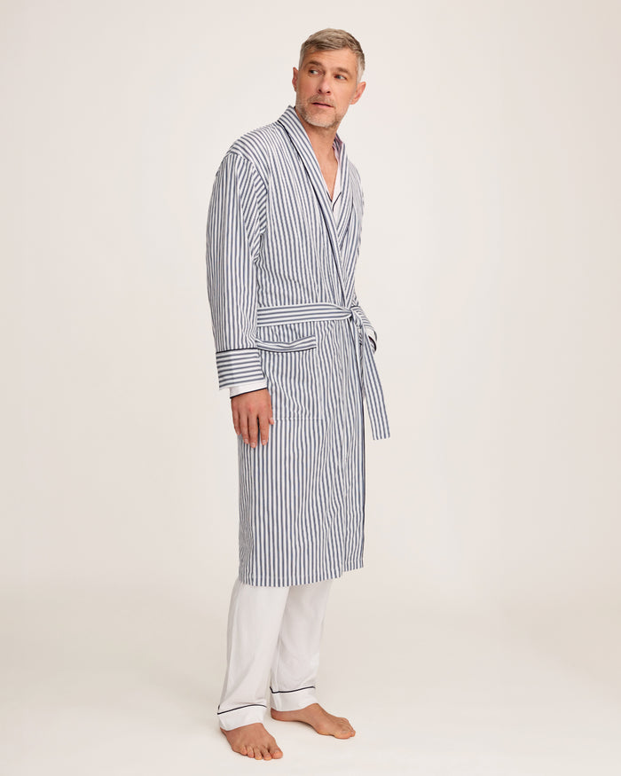 A man stands barefoot in white pajamas and the Men's Pima Robe in Navy French Ticking, belted, looking to the side against a plain light background.