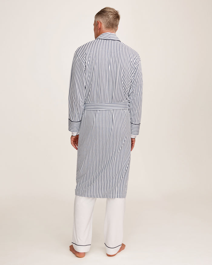 A man stands barefoot in white pajamas and the Men's Pima Robe in Navy French Ticking, belted, looking to the side against a plain light background.