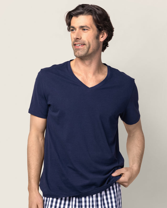 A man in a Mens Pima V-Neck Top in Navy by Petite Plume smiles and looks to the side, also wearing yarn-dyed checkered shorts against a plain, neutral background.