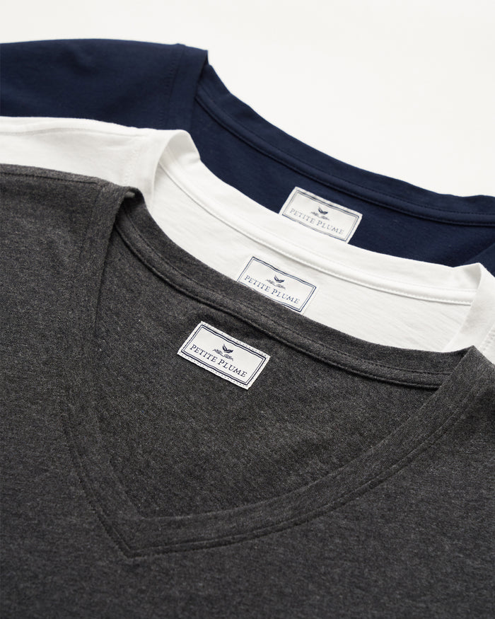 Three Petite Plume V-neck shirts, all made from Peruvian Pima cotton, are stacked: navy on top, white in the middle, and dark gray on the bottom. Each features a brand label near the collar.