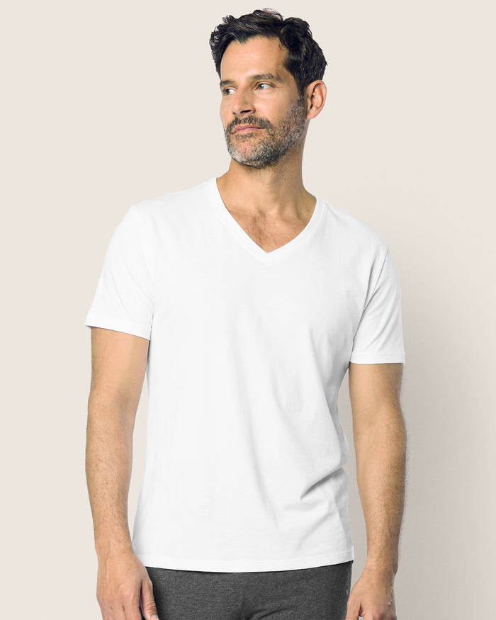 A man with short dark hair and a beard wears the Mens Pima V-Neck Top in White by Petite Plume, made from soft Peruvian Pima cotton. He stands against a neutral background, looking slightly to the side with a calm expression.