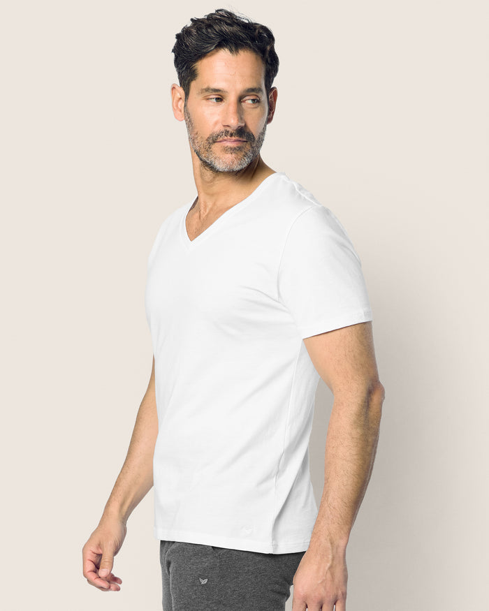 A man with short dark hair and a beard, wearing a Petite Plume Mens Pima V-Neck Top in White and dark pants, stands against a light grey background, looking to his left.