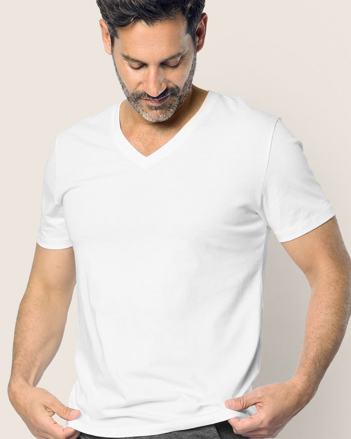 The man with short dark hair and a beard gazes down, donning the Mens Pima V-Neck Top in White by Petite Plume. He holds the sides of the shirt slightly outwards against a neutral beige background.