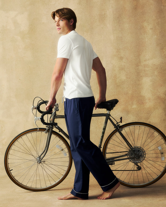 A barefoot man in a white shirt and Men’s Poplin Pants in Navy stands beside a vintage bicycle, looking over his shoulder against a tan textured background.