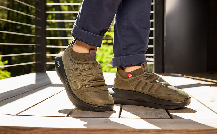 A person wearing olive green slip-on sneakers with dark gray soles, paired with navy blue pants, standing on a wooden deck outdoors | lifestyle