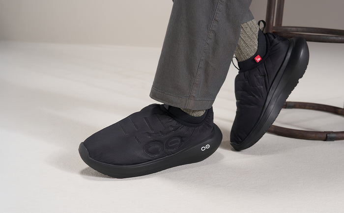 A person wearing black On running shoes and gray pants, shown from the calves down in a studio setting with a white floor and a dark chair leg visible in the background | lifestyle