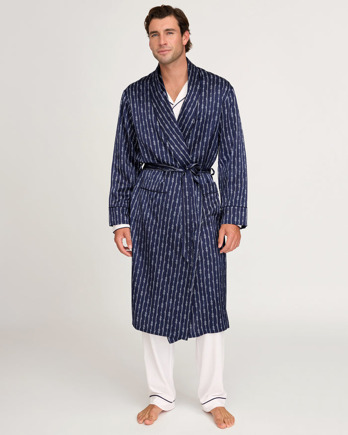 A man stands against a plain light background wearing the Men’s Silk Long Robe in Nautical Knots, tied at the waist over white pajamas with black piping.