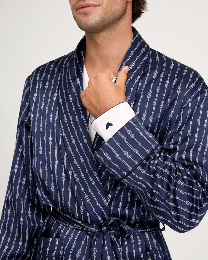 A man wears the Men’s Silk Long Robe in Nautical Knots, featuring a navy blue pattern with white lines, layered over a white dress shirt. He holds the robe’s lapel with one hand, displaying a cufflink on his shirt cuff against a plain, light-colored background.