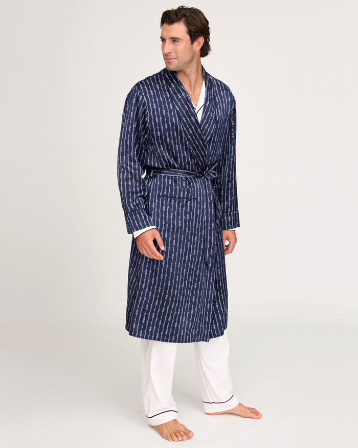 A man stands barefoot, smiling and looking to the side, wearing the Men’s Silk Long Robe in Nautical Knots over white pajamas with dark trim against a plain, light background.