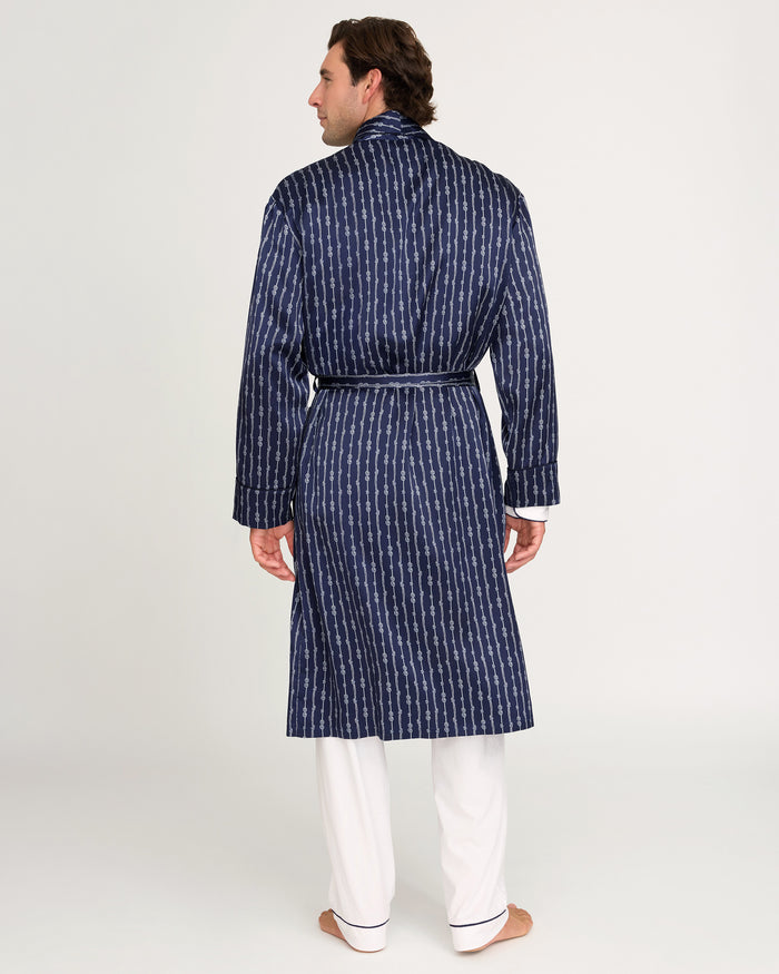 A man stands facing away, wearing the Men’s Silk Long Robe in Nautical Knots with a matching belt and white pajama pants. The background is plain and light-colored.