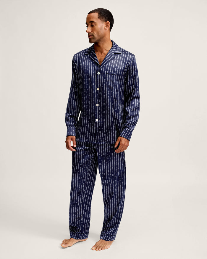 A man stands barefoot against a plain light background, wearing the Men’s Silk Pajama Set in Nautical Knots—a matching navy blue, long-sleeved, button-up pajama set with vertical white stripes. He is looking slightly to his left.