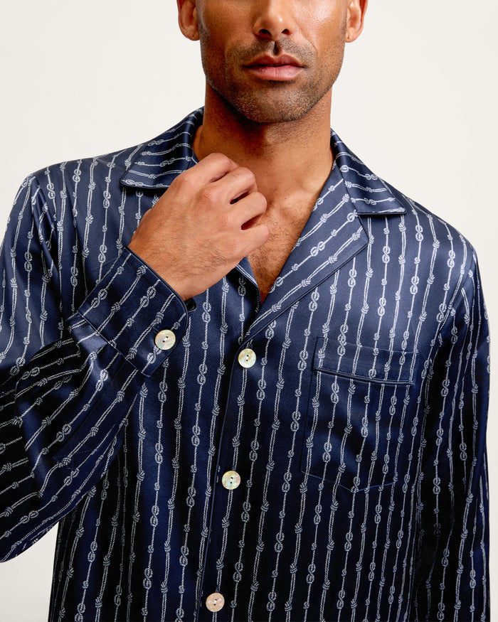 A man wearing the Men’s Silk Pajama Set in Nautical Knots—a navy silk pajama shirt with a white chain-link pattern—touches the collar with one hand. The shirt features a notched lapel and white buttons, set against a plain, light-colored background.