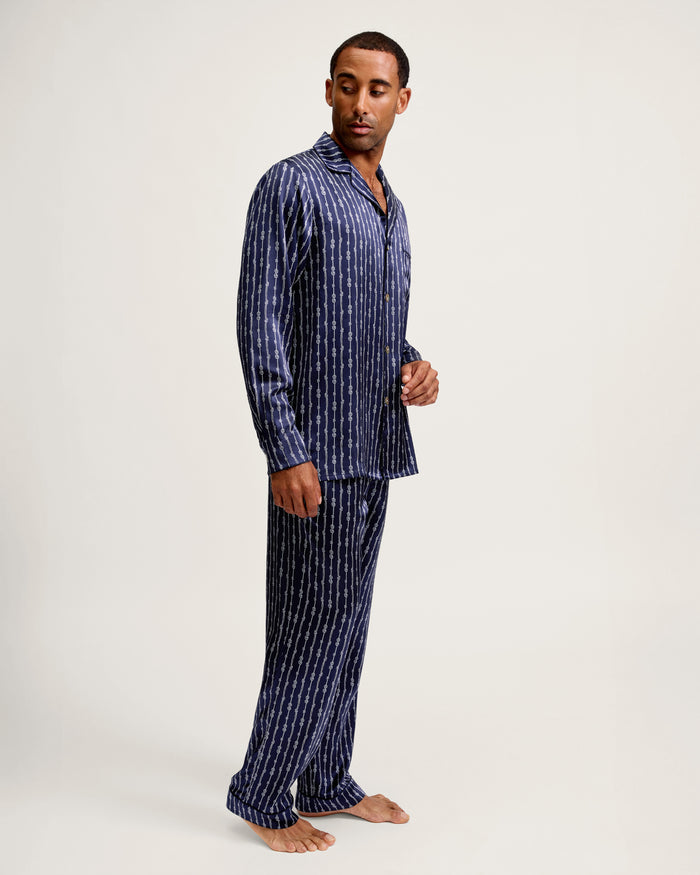 A man stands barefoot in the Men’s Silk Pajama Set in Nautical Knots, which features a navy blue base with vertical white stripes, gazing to the side against a plain light background.