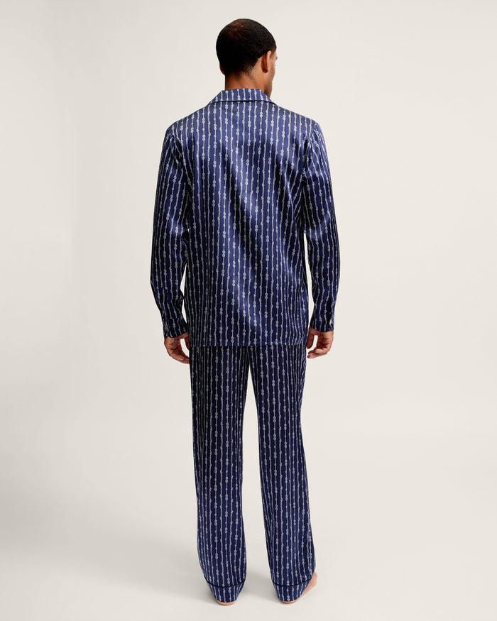 A person stands barefoot facing away, wearing the Men’s Silk Pajama Set in Nautical Knots—a matching dark blue patterned ensemble with long sleeves and pants—against a plain light background.
