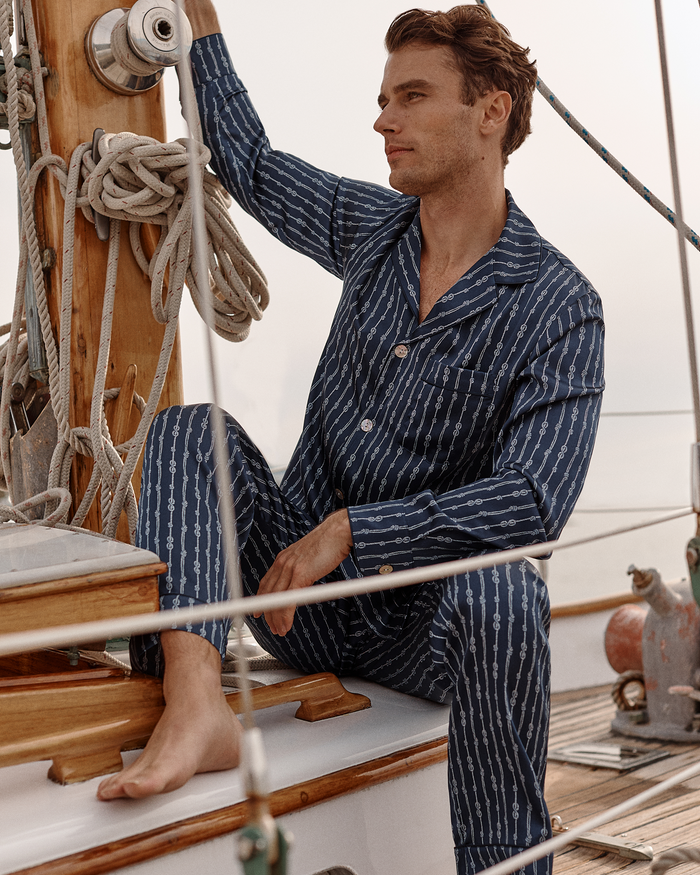A man wearing the Men’s Silk Pajama Set in Nautical Knots sits on a sailboat deck, gazing into the distance while holding a rigging line.