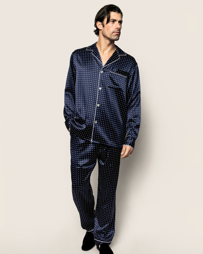 A man in the Petite Plume Mens Silk Pajama Set in Navy Polka Dot, featuring mulberry silk with white polka dots, stands against a plain beige background. He wears a button-up shirt and matching pants, has dark hair, and looks to his left.