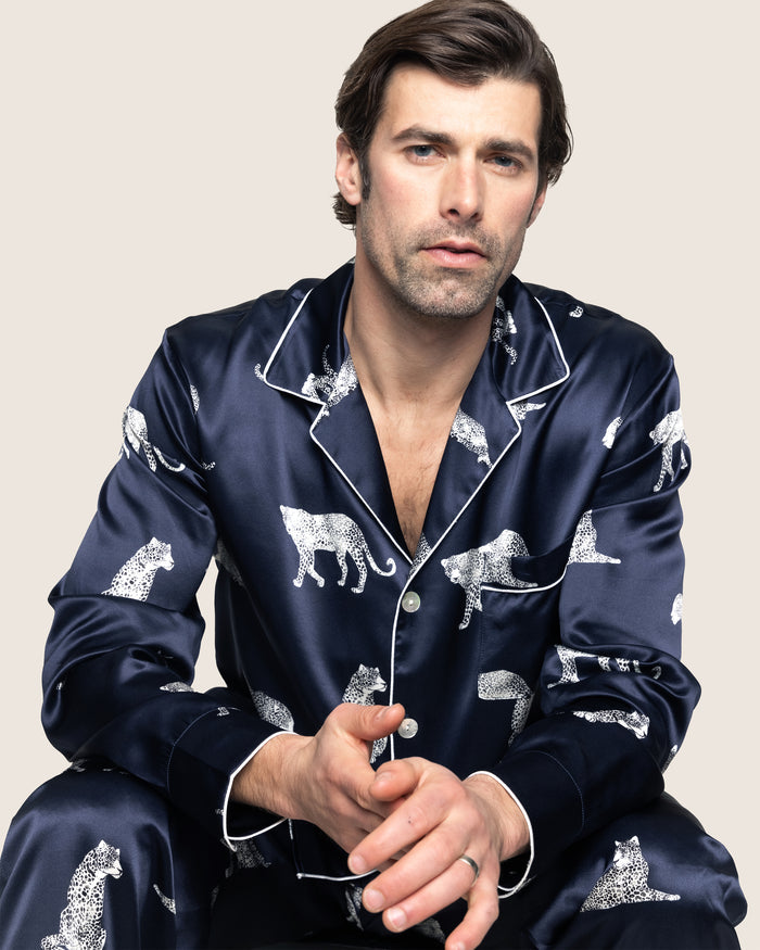 A man with dark hair and light facial hair sits looking forward, donning Petite Plumes Mens Silk Pajama Set in Panthère de Nuit, reminiscent of classic Hollywood style. The neutral solid background enhances the elegance of his silk sleepwear.