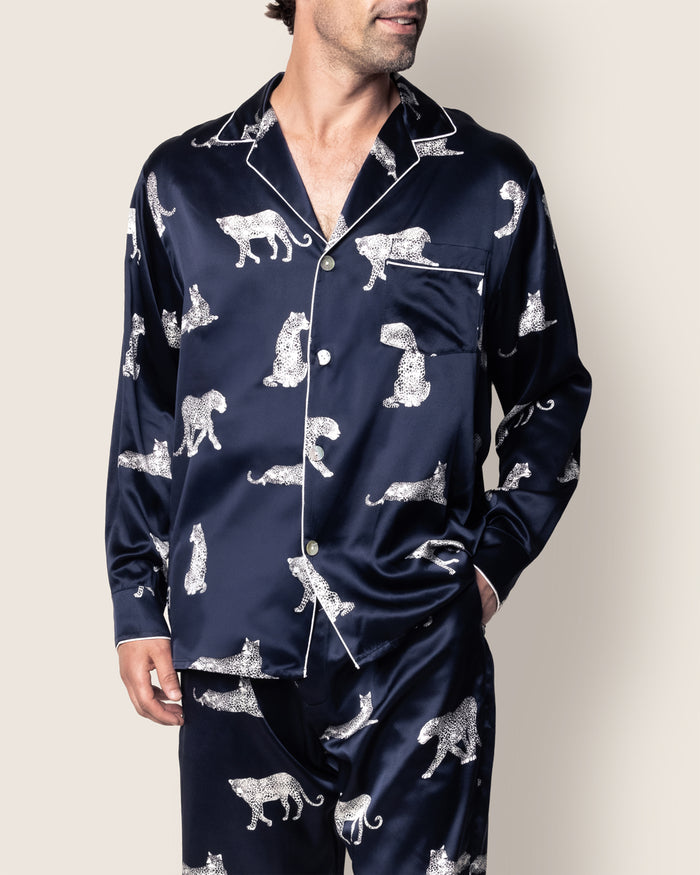 A person wearing the Mens Silk Pajama Set in Panthère de Nuit by Petite Plume exudes classic Hollywood elegance. The dark blue satin set, adorned with leopard illustrations, features a top with a front pocket and white piping, as they stand relaxed with hands partly in their pant pockets.